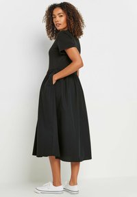 Vestido negro con un cuerpo ajustado y falda fluida, con mangas cortas, bolsillos laterales y una textura suave. Se lleva con zapatillas blancas.