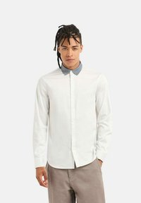 Camicia bianca a maniche lunghe con colletto a camicia grigio a contrasto, realizzata in tessuto liscio. Bottoni lungo la parte anteriore e polsini su misura.