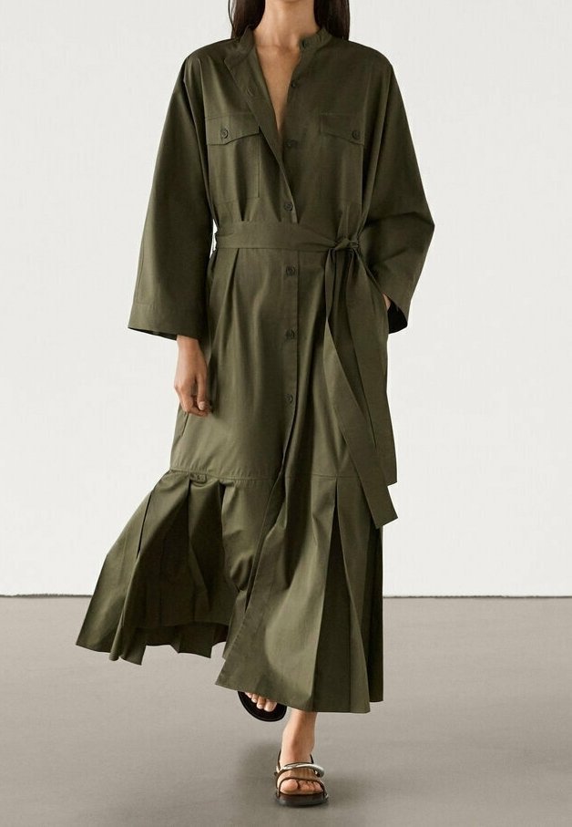 Femme portant une longue robe boutonnée vert olive avec une ceinture à la taille, des manches larges et un ourlet plissé, marchant en sandales ouvertes.
