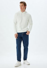 Witte capuchontrui met trekkoorden, gecombineerd met blauwe denim jeans en witte sneakers, met een relaxte pasvorm en een zachte textuur.