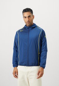 Blå vindjacka med gula detaljer och dragkedja framtill. Den har en huva och elastiska muddar, tillverkad av lätt material.