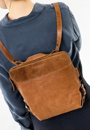 Persoon draagt een bruine leren rugzak over een donker, gestructureerd shirt met lange mouwen, gezien vanaf de achterkant en op schouderhoogte.