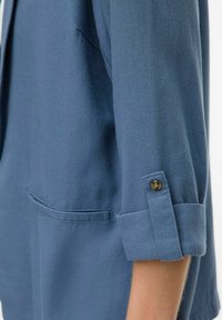 Mėlynas lininis švarkas su sulankstytu rankovu, turi mygtuko detalę ir priekinę kišenę. Minkšta tekstūra ir laisvas dizainas.