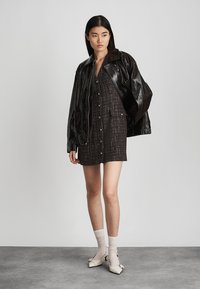 Abito camicia a quadri marrone con bottoni, abbinato a un giubbotto oversized nero lucido. La modella indossa calzini bianchi e scarpe color crema con cinturini.