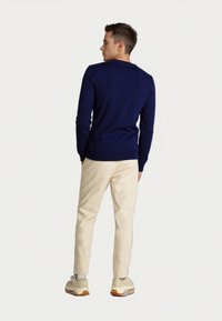 Hombre joven de pie mirando hacia un lado, vistiendo un suéter azul marino, pantalones beige, calcetines blancos y zapatillas beige con suelas de goma.