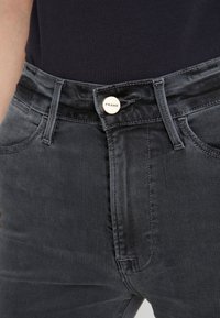 Jeans de denim gris con corte ajustado, que cuentan con un cierre de botón plateado etiquetado como "FRAME" y bolsillos frontales. Textura suave y diseño clásico.