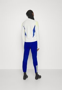 Bílý sportovní dlouhý rukáv s modrými a žlutými akcenty, doplněný o modré úzké kalhoty s bílým proužkovaným detailem po straně.