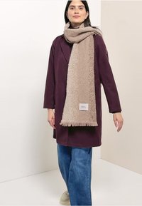Sciarpa beige texturizzata con dettagli sfrangiati drappeggiata su un cappotto viola, abbinata a jeans blu e scarpe chiare.