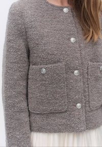 Harmaa tekstuuri takki pyöreällä kaula-aukolla, edessä nappikiinnitys ja kaksi päällyslaukkua, joissa on hopeanväriset napit ja pehmeä viimeistely.