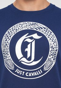 Laivyno mėlynos spalvos medvilninė marškinėliai su dideliu apvaliu sidabriniu grafiką, kuriame pavaizduota stilizuota "C", gyvatės ir tekstas "JUST CAVALLI."