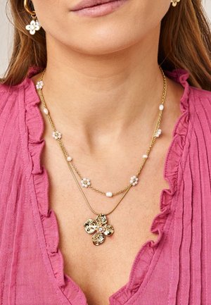 Mujer con collares dorados en capas, uno con pequeñas perlas blancas y flores, y otro con un gran colgante de flor dorada, combinados con pendientes a juego.