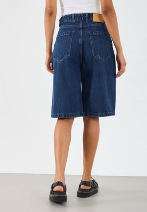 Frau trägt blaue Jeans-Shorts mittlerer Länge, weißes ärmelloses Oberteil und schwarze Sandalen mit Schnallen, steht mit dem Rücken zur Vorderseite.