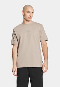 Beżowa koszulka z krótkim rękawem wykonana z miękkiej bawełny, z subtelną naszywką z logo z przodu, okrągłym dekoltem i luźnym krojem.