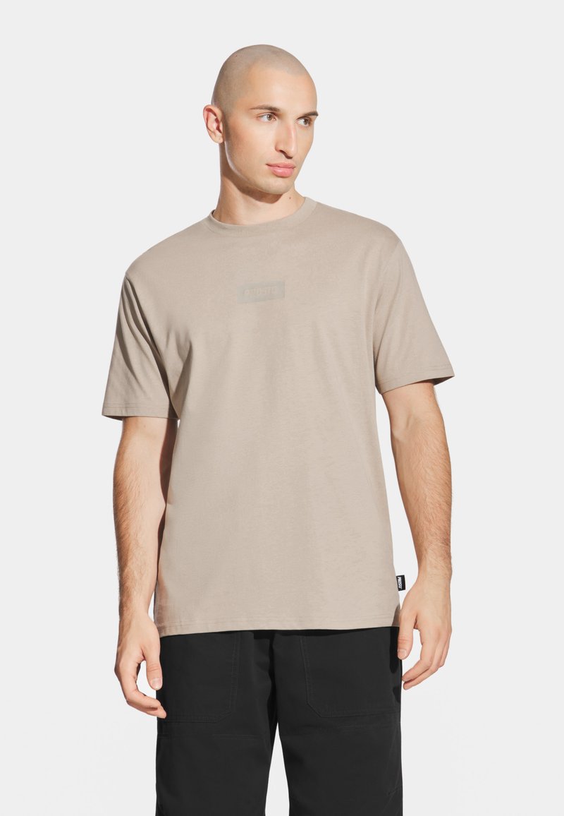 Beżowa koszulka z krótkim rękawem wykonana z miękkiej bawełny, z subtelną naszywką z logo z przodu, okrągłym dekoltem i luźnym krojem.