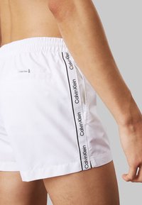 Shorts da bagno bianchi con cintura elastica, strisce laterali con il marchio "Calvin Klein" e una tasca sul retro. Texture morbida e taglio a lunghezza ginocchio.