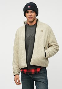 Muškarac nosi crnu pletenu kapu, bež flis jaknu preko sive dukserice i crveno-crnu kariranu košulju, stoji s jednom rukom u džepu jakne.