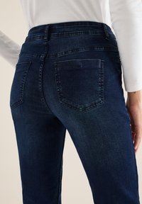 Tummansiniset farkut korkean vyötärön kanssa, joissa on kaksi takataskua ja sileä pinta, kuvattu sivusta.