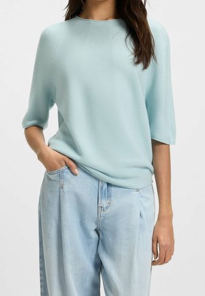 Frau trägt ein hellblaues, kurzärmeliges Strickoberteil und lockere hellblaue Jeans, mit einer Hand in der Tasche und der anderen entspannt.