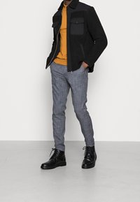 Černá fleecová bunda se šedými detaily, hořčicový svetr a šedé kárované kalhoty. Černé šněrovací boty doplňují outfit.