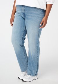 Personne portant un jean droit bleu clair et des baskets blanches, se tenant devant un fond blanc uni.