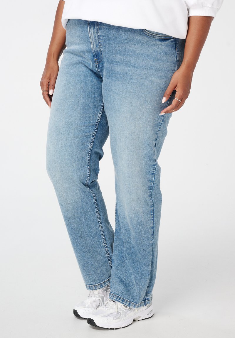 Personne portant un jean droit bleu clair et des baskets blanches, se tenant devant un fond blanc uni.