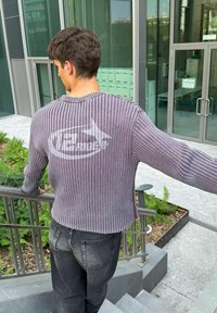 Grauer gerippter Pullover mit langen Ärmeln und abgerundetem Saum, trägt ein weißes Logo auf der Rückseite. Kombiniert mit dunklen Jeans.