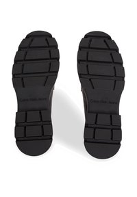 Suole di scarpe in gomma nera con un motivo testurizzato. Presenta il logo "Calvin Klein Jeans" vicino al tallone.