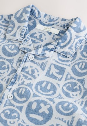 Chemise à manches courtes avec col en tissu bleu clair présentant divers motifs de smileys et d'icônes, avec une fermeture boutonnée sur le devant et une texture douce.