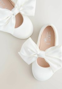 Weiße Ballerinas mit einer großen, strukturierten Satinschleife auf der Vorderseite. Glatte Oberfläche, abgerundete Spitze und gepolsterte Innensohle.