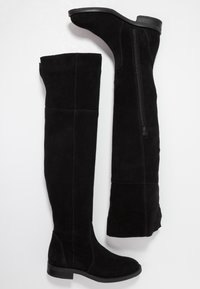 Bottes noires en daim au-dessus du genou avec semelle plate, fermeture éclair sur un côté et détails de coutures verticales. Texture douce et design élégant.