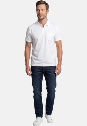 Uomo con polo a maniche corte bianca con tasca con cerniera, jeans blu scuro e sneakers bianche, in piedi e che guarda a destra.