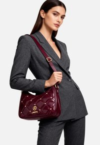 Borsa a tracolla in pelle verniciata bordeaux con motivo trapuntato, dettagli in metallo dorato e logo decorativo. Tracolla regolabile per una portabilità versatile.