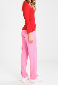 Rode langemouwen shirt met uitlopende manchetten, gecombineerd met roze wijde broek. Beide zijn effen van kleur, gemaakt van een gladde stof, gedragen met witte sneakers.
