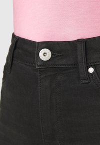 Svarta denimjeans med en silverknapp prydd med "M&S EST 1884", med midja i normalhöjd och rak passform. Bakgrunden visar en rosa topp.