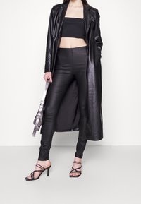 Černý kožený dlouhý kabát přes černý crop top, doplněný lesklými černými přiléhavými kalhotami a sandály na vysokém podpatku se šněrováním. Stříbrná taška jako akcent.