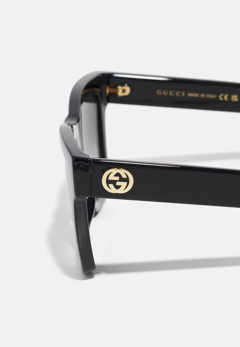 Lunettes de soleil rectangulaires noires avec finition brillante. Présentent un détail de logo doré sur le côté et des montures en plastique robustes. Les verres sont teintés foncés.