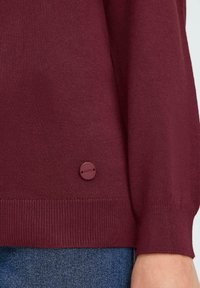 Maglione lavorato a maglia color bordeaux con polsini a coste, caratterizzato da un piccolo badge circolare sul bordo. Texture morbida, vestibilità comoda e colore uniforme.