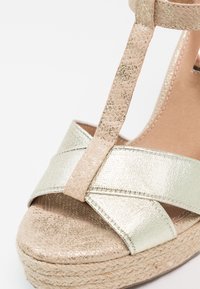 Sandale à plateforme avec un design de bride croisée en métal doré, une bride texturée beige et une semelle en espadrille tissée. Fini lisse et tons neutres.