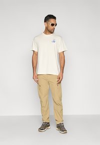 Μπεζ βαμβακερό t-shirt με ανοιχτό μπλε λογότυπο, συνδυασμένο με χακί παντελόνι cargo με τσέπες με φερμουάρ και καφέ παπούτσια πεζοπορίας με μαύρες λεπτομέρειες.