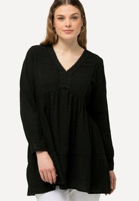 Zwarte tuniek met lange mouwen, V-hals en textuurstof. Kenmerkend zijn de decoratieve kantafwerking en de gelaagde zoom.