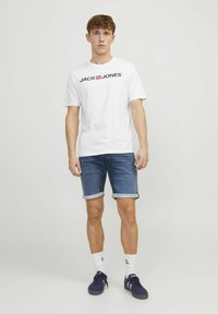 T-shirt bianca a maniche corte con logo "JACK & JONES", abbinata a pantaloni corti di jeans blu con risvolti e sneakers navy con calzini bianchi.