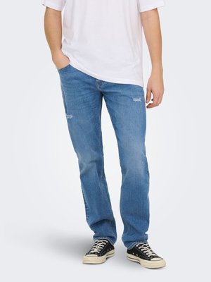 Persoon draagt lichtblauwe spijkerbroek met lichte slijtage, wit T-shirt en zwarte sneakers met lage schacht en witte veters.