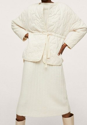 Personne portant une veste matelassée crème avec fermeture par lien sur une robe en tricot côtelé et des bottes claires, debout les mains sur les hanches.