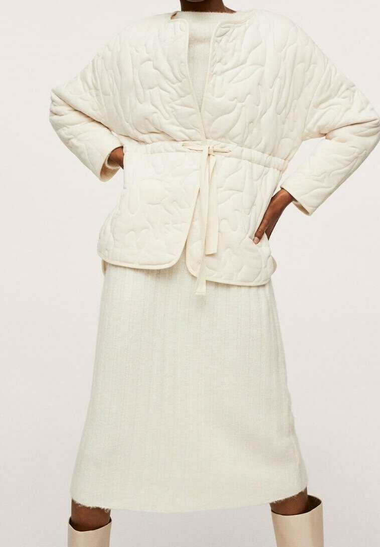 Personne portant une veste matelassée crème avec fermeture par lien sur une robe en tricot côtelé et des bottes claires, debout les mains sur les hanches.