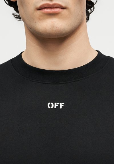 Άνδρας που φοράει μαύρο πουκάμισο με λευκό κείμενο "OFF" στο κέντρο κάτω από το λαιμό, σε απλό φόντο.