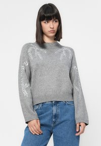 Grå genser laget av mykt materiale, med broderte bladmønstre på ermene, ribbestrikket halslinning og en avslappet passform. Kombinert med blå jeans.