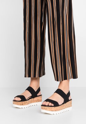 Sandali platform neri con intersuola in sughero, caratterizzati da ampie fasce nere e suola bianca texturizzata, indossati con pantaloni a righe verticali.