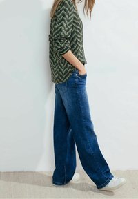 Chemise verte à manches longues avec un motif en zigzag, associée à un jean bleu ample et des baskets blanches. Fond de mur avec une texture légère en bas.