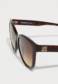 Óculos de sol em padrão tartaruga com uma armação ousada e angular. Lentes marrons em gradiente, marcação em dourado na haste e textura plástica suave.