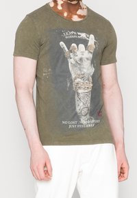 T-shirt vert olive, col rond, avec un graphique représentant une main avec des motifs de bagues et du texte. Le tissu a une légère texture. Porté avec un pantalon blanc.